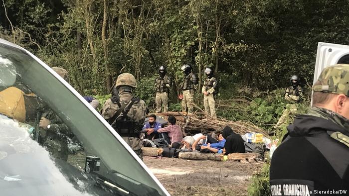 Border police guarding a group of migrants sitting in a wooded area