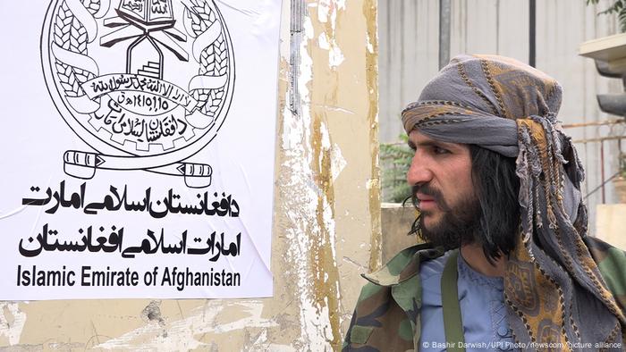 Un combattant taliban à Kaboul le 17 août 2021 à côté d'une affiche proclamant « l'Émirat islamique d'Afghanistan »