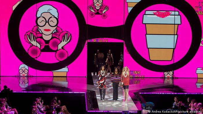 A fashion show with three models on the catwalk and a big pink display in the background reproducing Iris Apfel imagery.