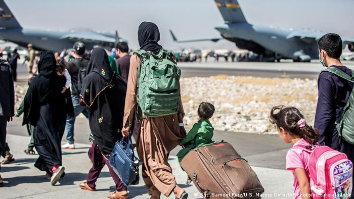 line of people with suitcases and backpacks, including children, walking on the Kabul airport tarmac toward planes