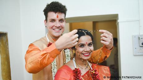 Ein Mann steht hinter einer Frau und legt ihre eine Kette um. Beide lächeln und tragen traditionell indische Kleidung.