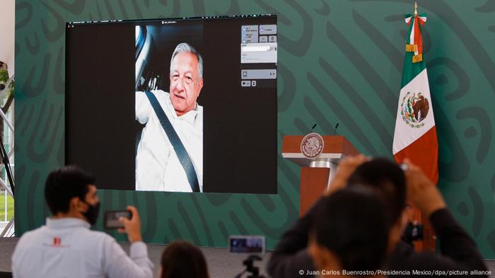 A screen in a conference room shows Lopez Obrador delivering his remarks via video call on his phone from a car