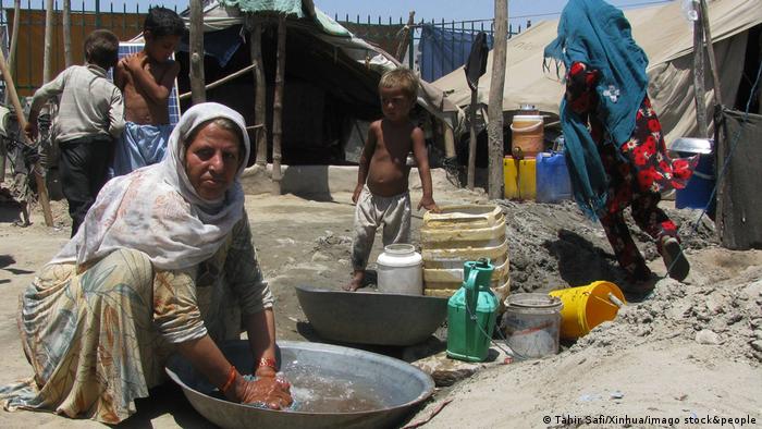 An Afghan woman wearing a headscarf washes her clothes at a refugee camp. A child stands in the background. 