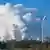 Water vapour pours from cooling towers, a wind turbine in the foreground