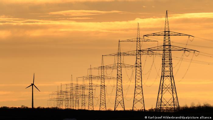 Eine Windkraftanlage steht neben Hochspannungsleitungen im Sonnenuntergang bei Seligenstadt in Bayern, Deutschland