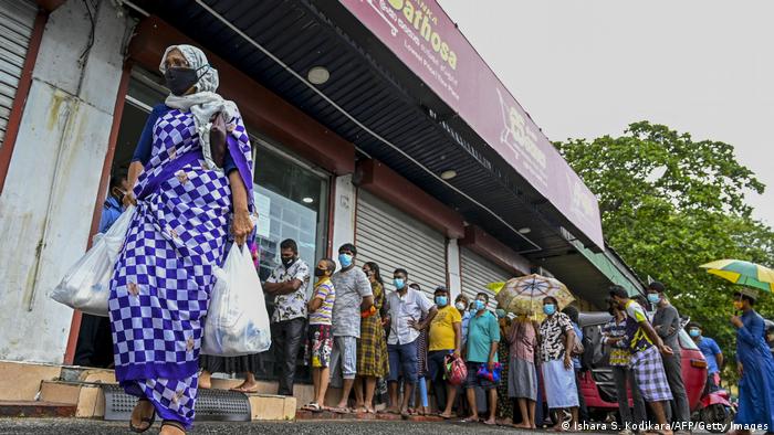 A woman carrying food bags walks pasts people standing in queue outside a state-run supermarket to buy essential food items in Colombo
