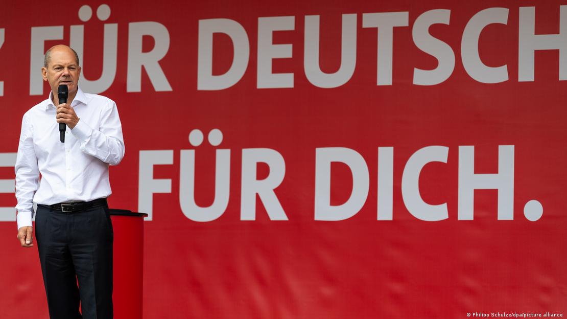 SPD-Kanzlerkandidat Olaf Scholz steht in einem weißen Hemd vor einem roten Plakat, auf dem in großen weißen Buchstaben "Für Deutschland, für Dich" steht. Er hält ein Mikrofon in der Hand und spricht. SPD-Kanzlerkandidat Olaf Scholz steht in einem weißen Hemd vor einem roten Plakat, auf dem in großen weißen Buchstaben "Für Deutschland, für Dich" steht. Er hält ein Mikrofon in der Hand und spricht.