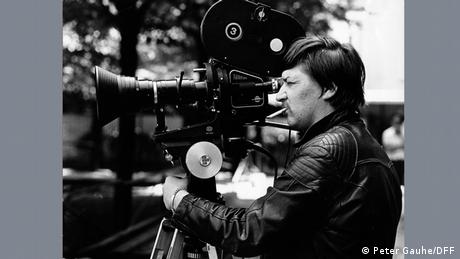 Fassbinder , a man in a black leather jacket, cigarettwe dangling from his lips, stands looking into a big camera