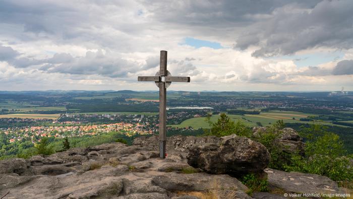 Ein monumentales Kreuz über einem Hügel überblickt Corlitz in einem Tal unter einem bewölkten Himmel