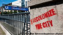 Vor dem Eingang der U-Bahnstation Mohrenstraße hat jemand mit roter Farbe decolonize the city an die Wand geschrieben.