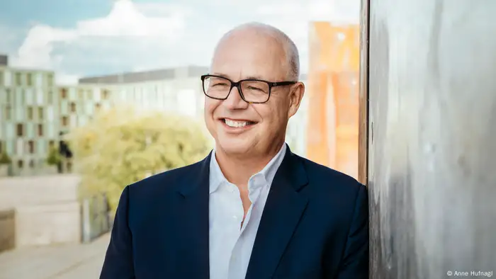 Sven Gösmann smiles in front of a blurred city background wearing a blue jacket and white open-collar shirt.