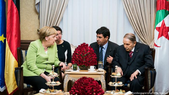 Merkel und Bouteflika und weitere Personen sitzen an einem geschmückten Tisch