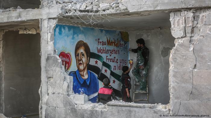 Man standing on a ladder painting a wall of a destroyed building. He is assisted by another man and watched by a boy.