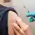 Hand in blue plastic glove holding a vaccination needle next to a bared upper arm