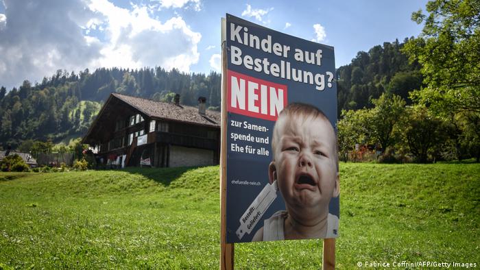 Paisagem idílica com chalé, em 1º plano, cartaz com bebê chorando e legendaCrianças por encomenda? Não, em alemão