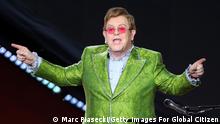 Elton John in a green suit pointing his index fingers up.