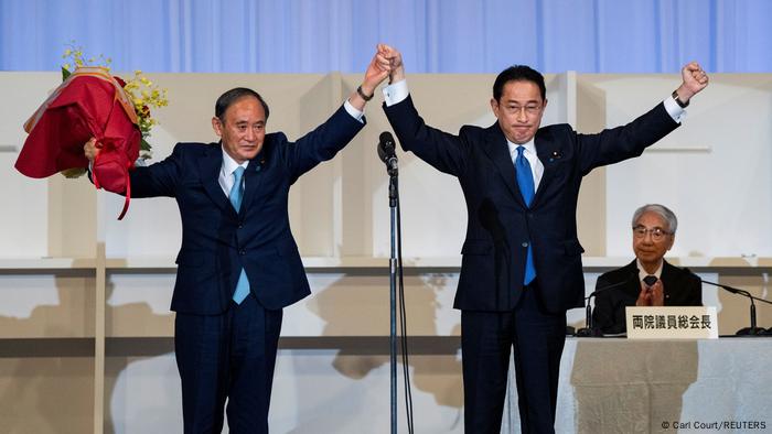 Fumio Kishida (r.) und Yoshihide Suga (l.) stehen auf einer Bühne und halten triumphierend die Fäuste in die Luft, Yoshihinde Suga hält dabei einen Blumenstrauß in der linken Hand und die Hand Kishidas in seiner rechten. Fumio Kishida (r.) und Yoshihide Suga (l.) stehen auf einer Bühne und halten triumphierend die Fäuste in die Luft, Yoshihinde Suga hält dabei einen Blumenstrauß in der linken Hand und die Hand Kishidas in seiner rechten.