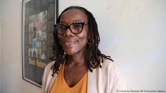 Tsitsi Dangarembga: Woman smiles into camera, poster on a wall in the background