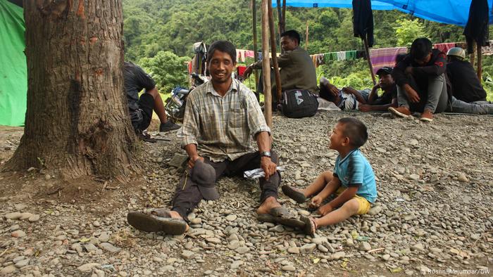 A man and his son are sitting smiling on the ground next to a tree, several other men are sitting behind them A man and his son are sitting smiling on the ground next to a tree, several other men are sitting behind them
