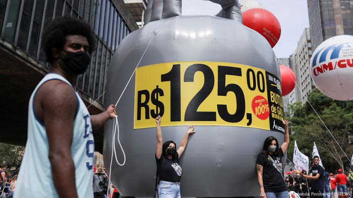Homem ao lado de botijão de gás gigante com o preço de R$ 125