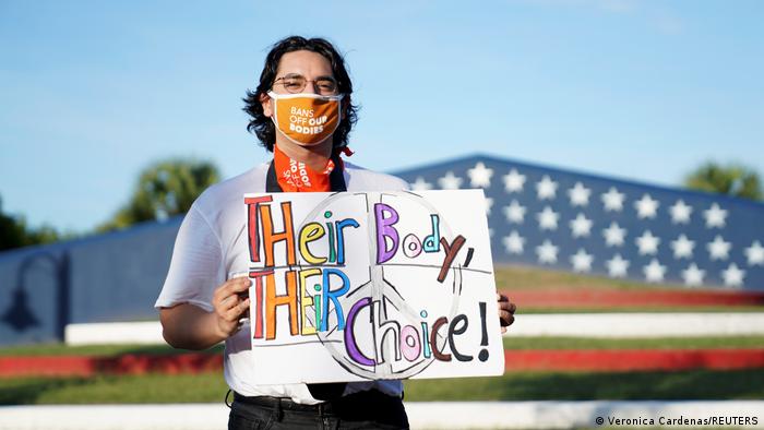 Foto de una persona que sostiene un rótulo en inglés que dice su cuerpo, su decisión frente a una bandera de EE.UU.
