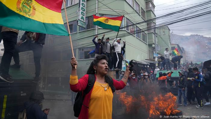 Protesta de cocaleros en La Paz: una mujer y varios grupos de manigestantes alzan banderas de Bolivia en medio de una calle en la que arden neumáticos, frente a la sede de ADEPCOCA