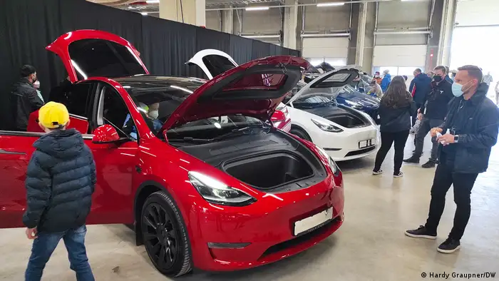 Visitors examine red Tesla sedan with its hood, trunk and doors open, next to a white one