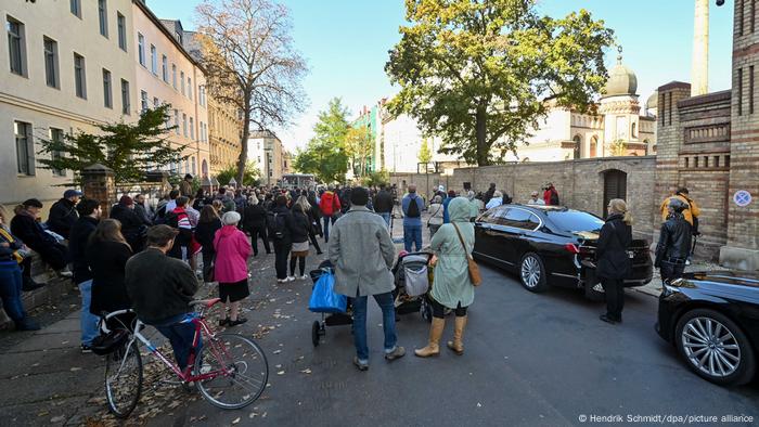 Blick in eine Straße, in der viele Menschen zusammengekommen sind und nachdenklich innehalten, rechts steht hinter einer Mauer ein Gebäude mit einem Turm 