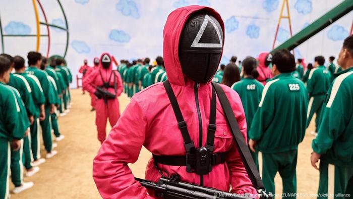 A figure wearing a red suit and a mask points a gun at people standing in line