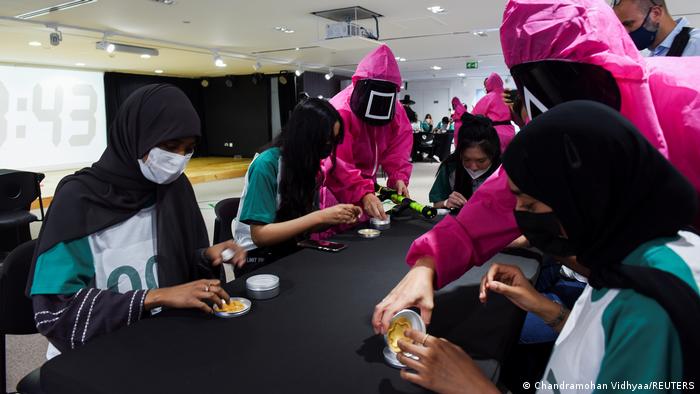 Squid Game Challenge : des personnes portant des foulards et des masques sont assises autour de la table, des personnes portant des costumes roses se tiennent à côté d'elles