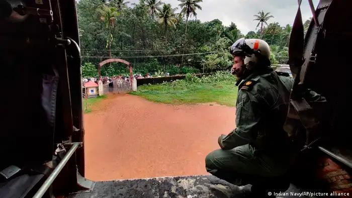 a soldier looks out from a helicopter at a group of people waiting for relief with a red swollen river in front of them