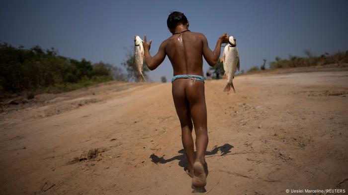 Um jovem indígena de costas, com um peixe em cada mão 