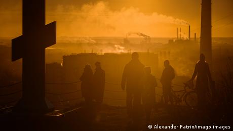 People walk under an orange sky in the city of Kemerovo, Russia, next to the silhouette of a cross, as smog hangs over the city in the background and factories with smokestacks are seen in the distance