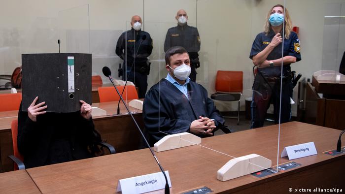 A woman seated in a courtroom covers her face behind an open folder as she sits next to a lawyer and is watched by three police officers
