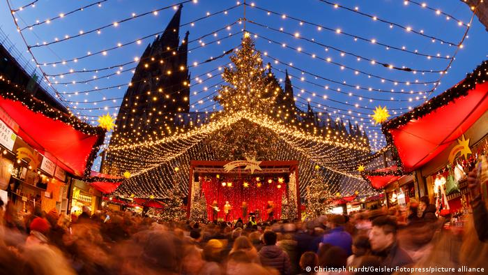 Festive lights over a Christmas market create a roof over people watching the entertainment on a stage next to Cologne Cathedral