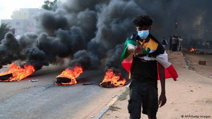 A masked Sudanese protester signalling victory as tires burn in the background 