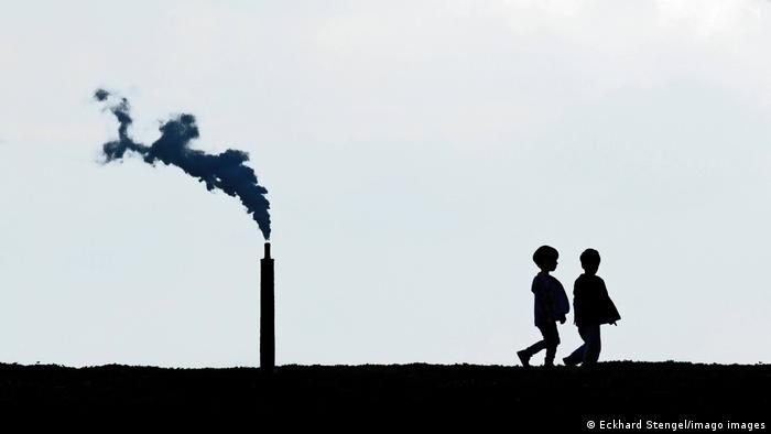 Zwei Kinder gehen auf einem Deich spazieren. Im Hintergrund der Schornstein einer Chemiefabrik, die Titandioxid hergestellt.