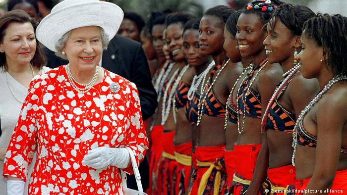 Queen Elizabeth wears a colorful red dress standing next to dancers from Mozambique.