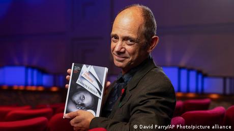 Porträtfoto von Damon Galgut, er guckt seitlich in die Kamera, in der Hand hält er eine Ausgabe seines Buches The Promise