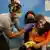 Menino de máscara colorida está sentado no colo da mãe. Uma mulher de luvas azuis aplica a vacina no braço dele. Ele faz cara de susto. A mãe está de olhos fechados. 