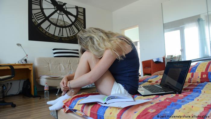 Девушка сидит на кровати в своей комнате, опустив голову на колени