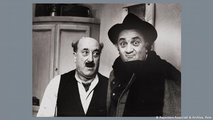 Setfoto: Federico Fellini steht mit Hut, Mantel und Schal neben Armando Brancia und zieht eine Grimasse.