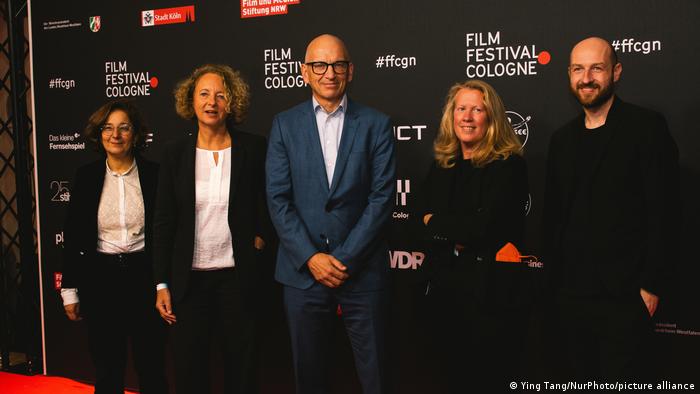 Direktor Phil Grabsky (Mitte) steht zwischen Mitarbeitenden des WDR vor einem Plakat des Film Festivals Cologne 