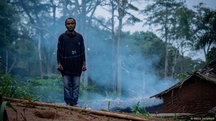 A man stands as smoke from his hut wafts into the forest behind him