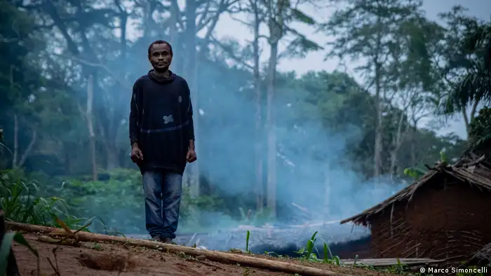A man stands as smoke from his hut wafts into the forest behind him
