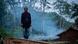 A man stands as smoke from his hut wafts into the forest behind him