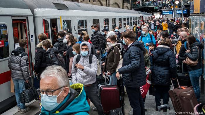Sebuah platform yang ramai di Hamburg;  orang-orang dengan topeng dan koper menunggu pintu kereta dibuka.