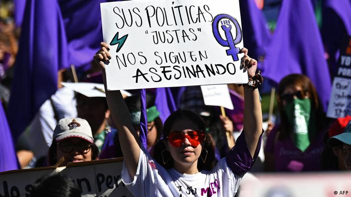 Foto simbólica de una mujer que protesta en El Salvador con un rótulo que dice sus políticas justas nos siguen asesinando.