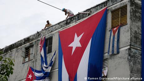Unos hombres cuelgan banderas cubanas sobre las ventanas de la casa del activista opositor Yunior García Aguilera en un intento de impedir que se comunique con el exterior, mientras sostiene una flor desde una ventana.