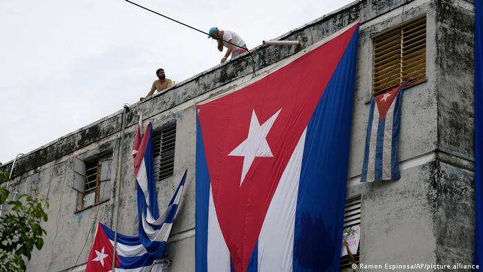 Men on a rooftop lower massive Cuban flags over the facade and windows of a building